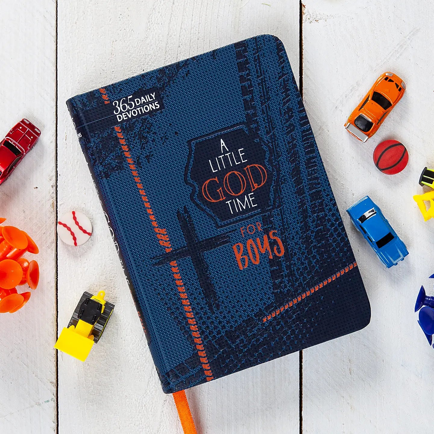 A blue faux leather devotional book titled 'A Little God Time for Boys' placed among children's toy cars and a basketball on a white wooden surface.
