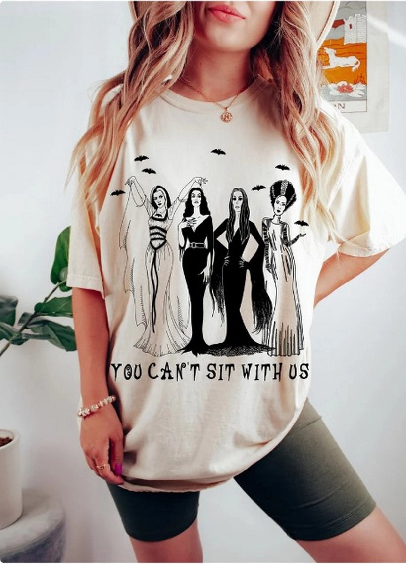 Woman wearing a cream t-shirt with a graphic of the Women of Halloween that says 'You Can't Sit With Us'