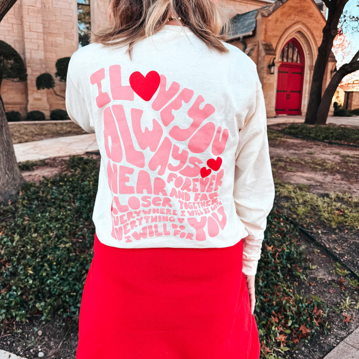 A woman wearing a cream long sleeve graphic tee with a heart design on the pocket and a love message on the back, that reads 'I love you always, forever, near and far closer together everywhere I will be everything I will do for you.'