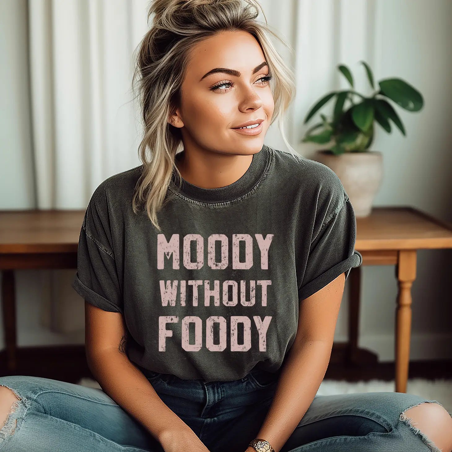 A woman wearing a gray graphic tee that says 'MOODY WITHOUT FOODY' paired with ripped jeans.