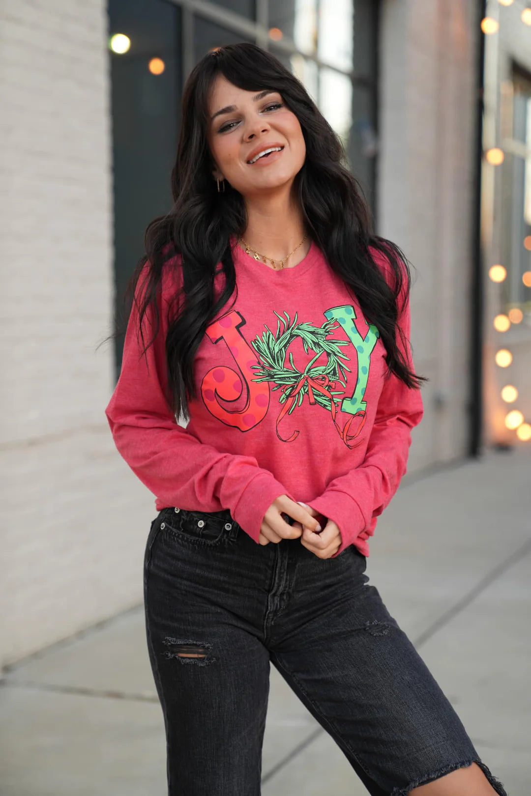 A woman wearing a red long sleeve graphic tee with the word 'JOY' printed on the front with Christmas graphics.