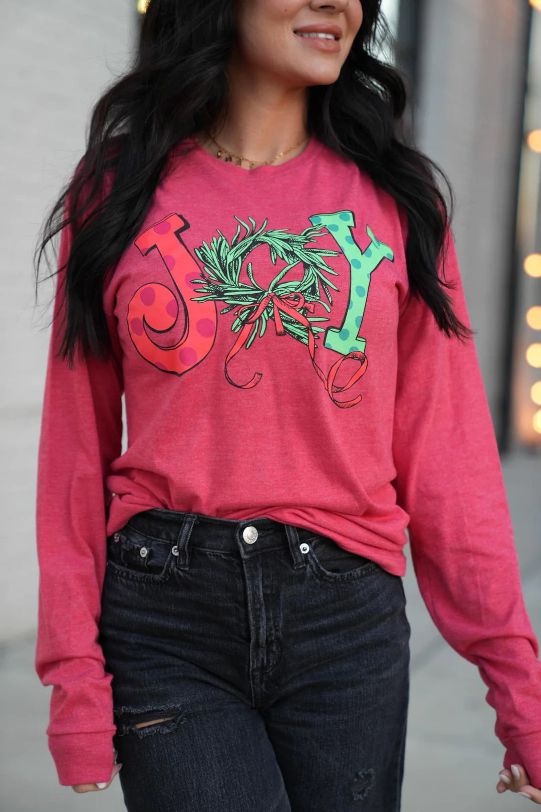 A woman wearing a red long sleeve graphic tee with the word 'JOY' printed on the front with Christmas graphics.