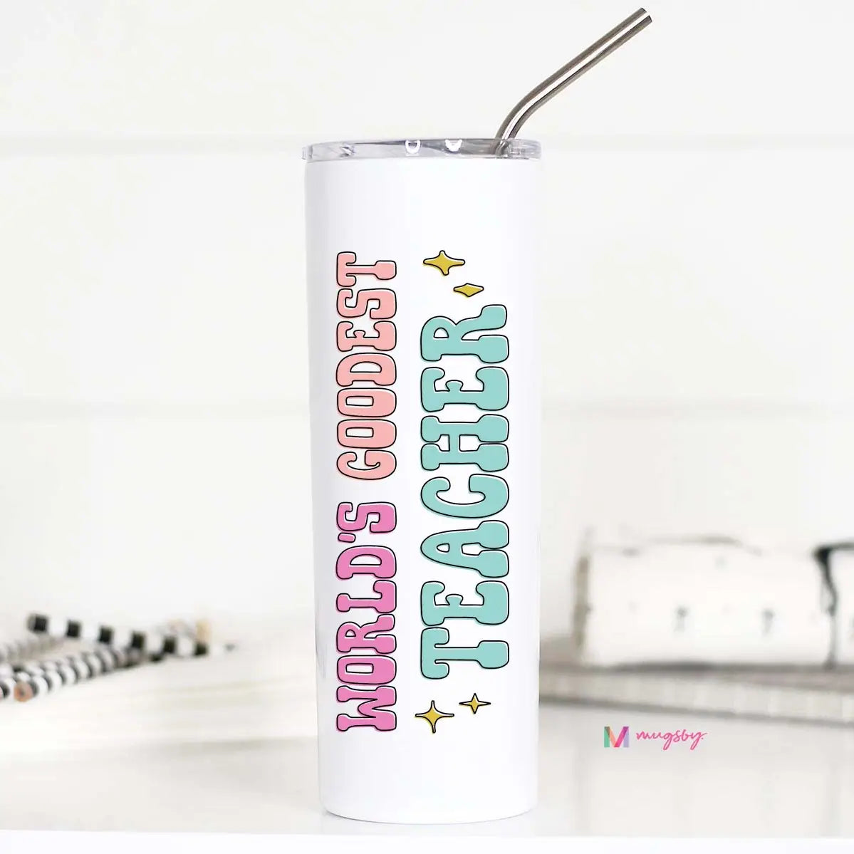 A tall stainless steel travel cup with a 'World's Goodest Teacher' print on it, featuring a metal straw and plastic lid, on a desk alongside a notebook and pens.