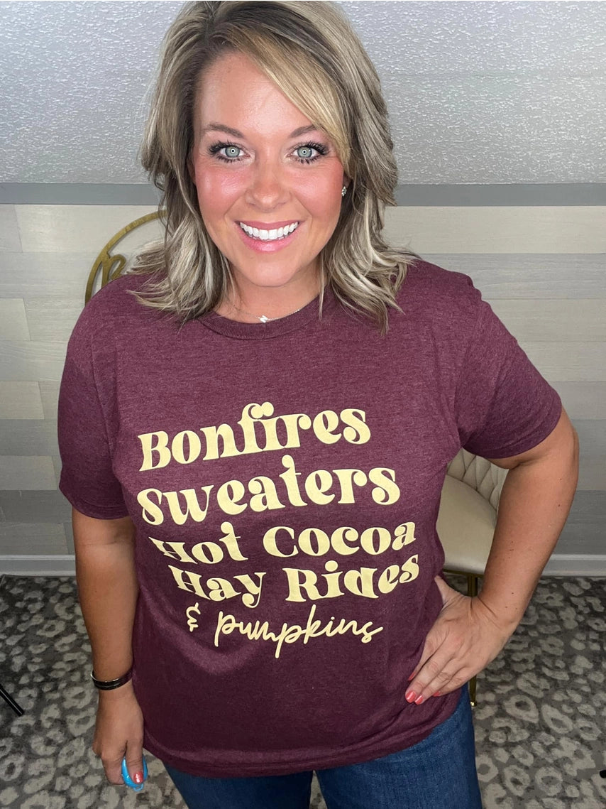 Woman wearing a maroon t-shirt that reads,  'Bonfires, Sweaters, Hot cocoa, Hay Rides and Pumpkins.' written in a cream text.