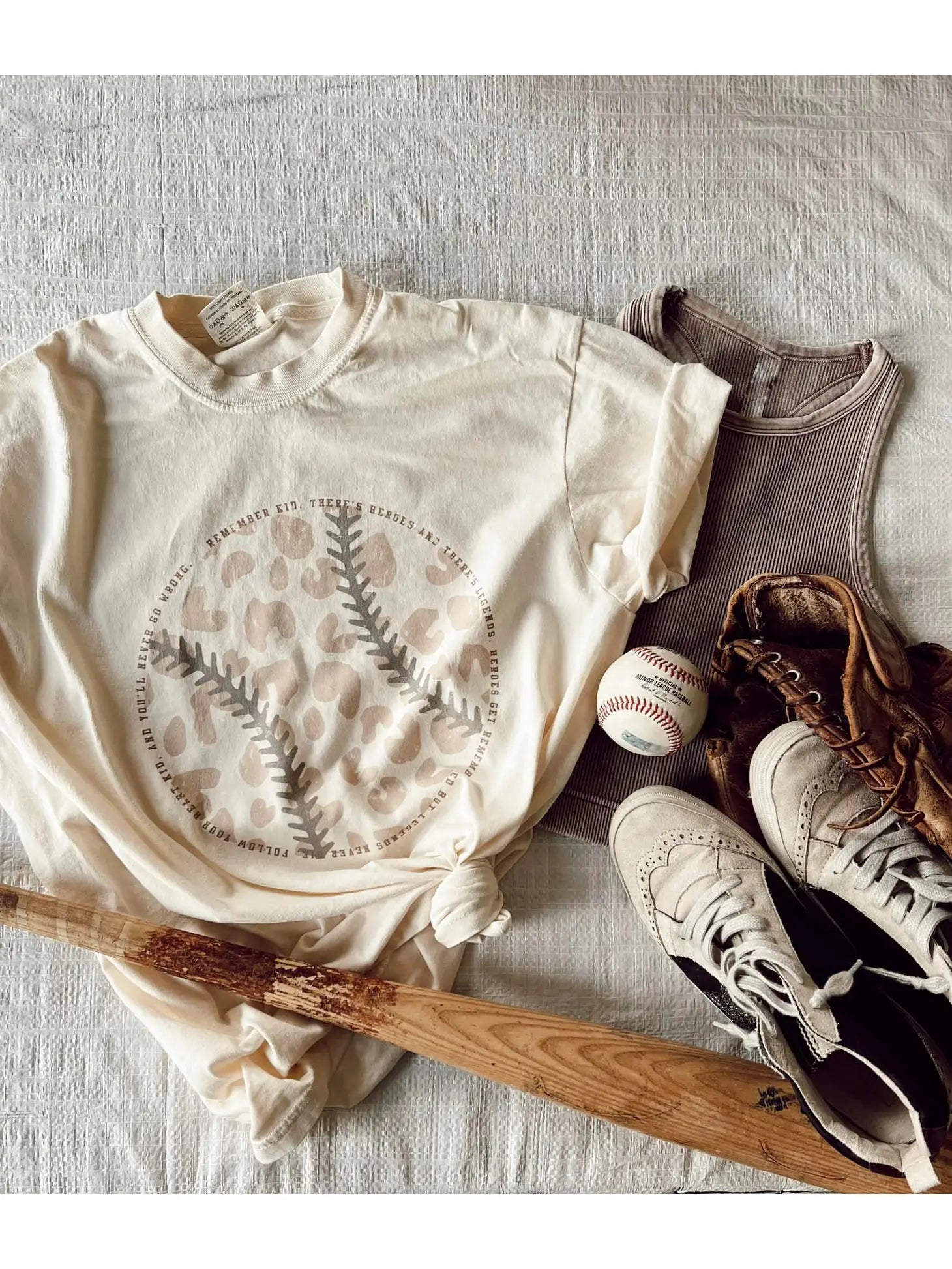 A cream baseball graphic tee with a pattern resembling a baseball and text, 'Remember kid, There's heroes and there's legends. Heroes get remembered but legends never die. Follow your heart kid, and you'll never go wrong.'