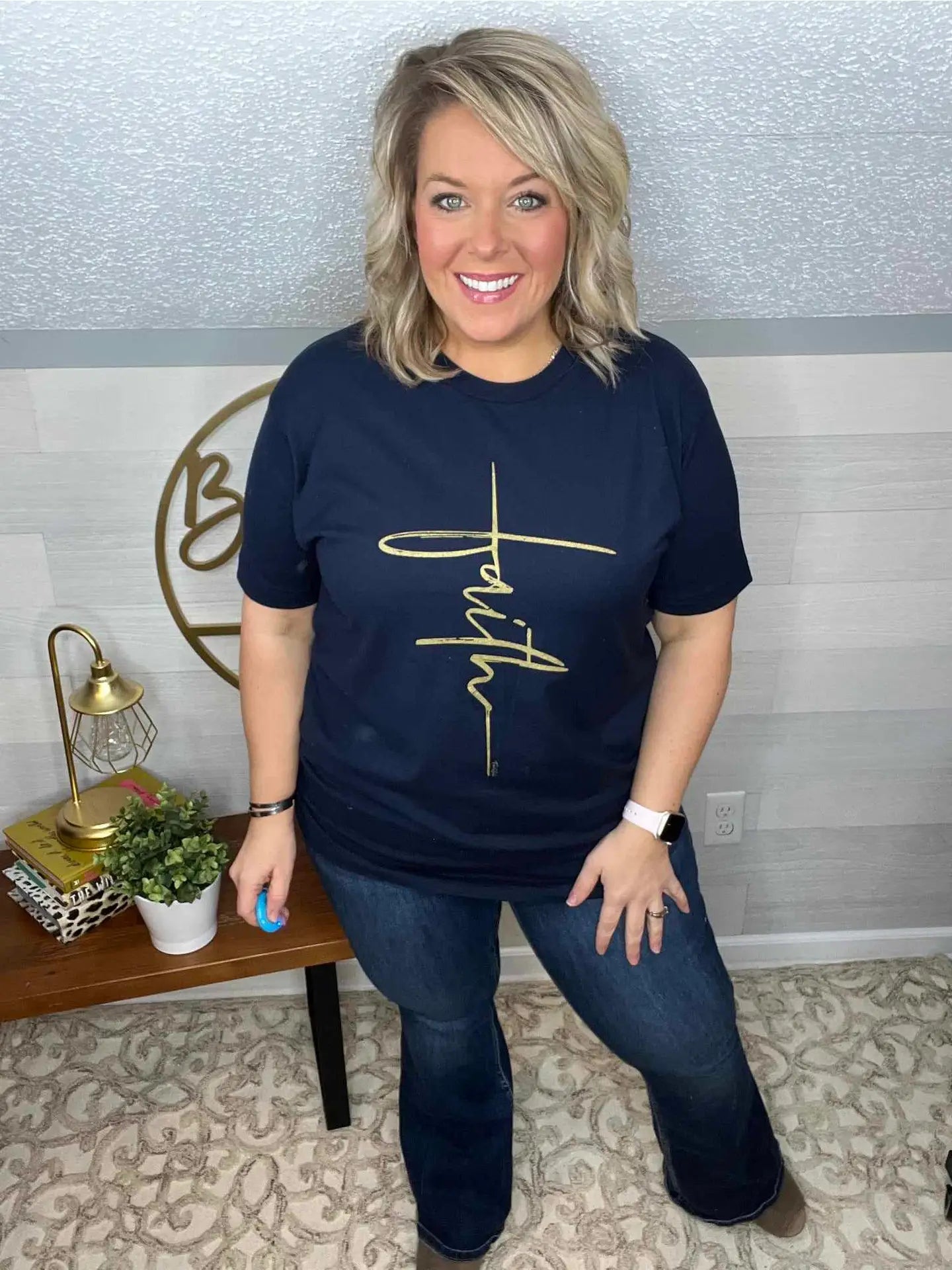 A woman wearing a navy crew neck t-shirt with a vertical gold cross graphic with the word 'Faith' written in it.