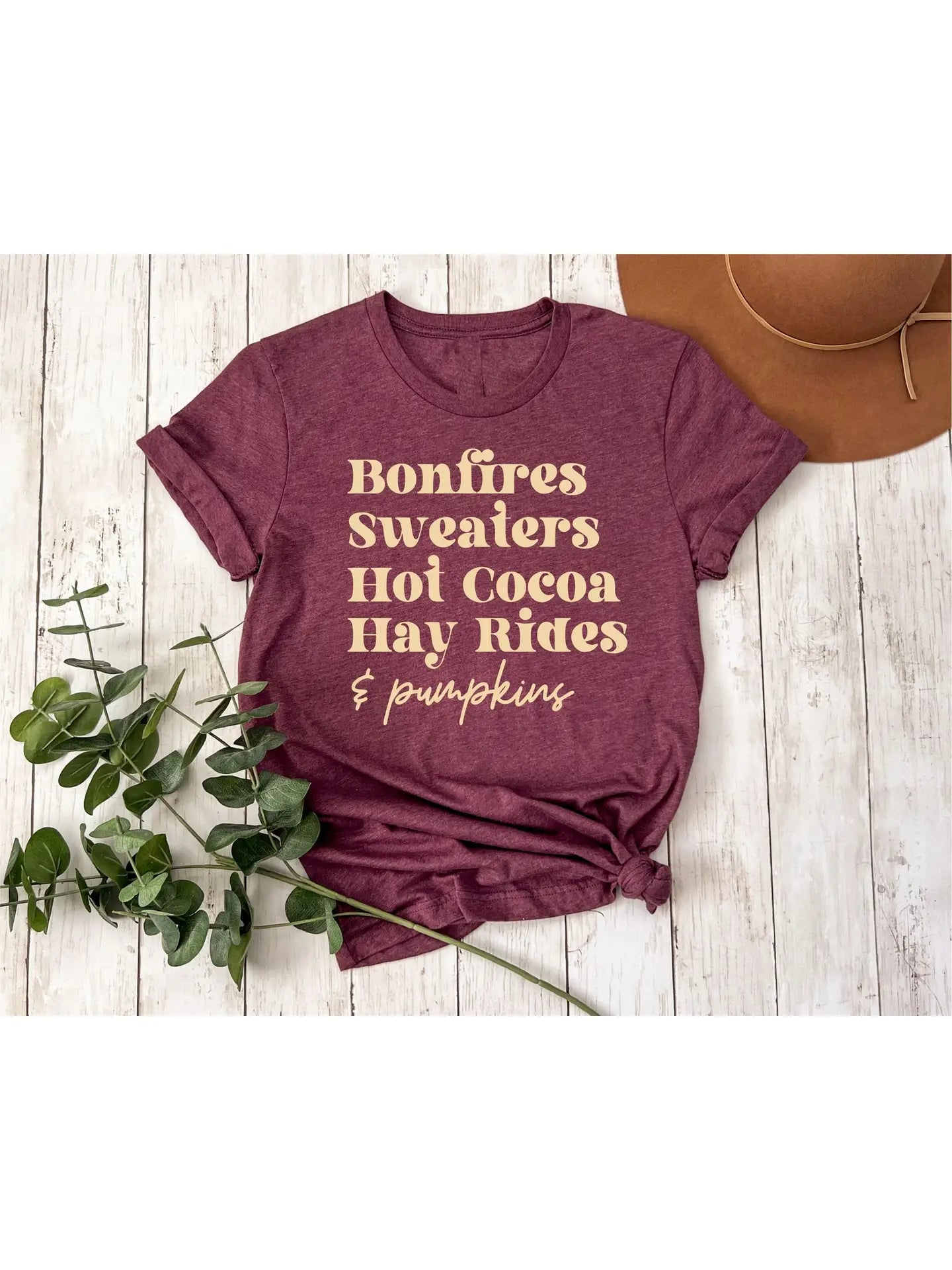 A maroon t-shirt that reads,  'Bonfires, Sweaters, Hot cocoa, Hay Rides and Pumpkins.' written in a cream text.