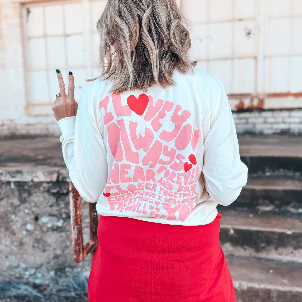 A woman wearing a cream long sleeve graphic tee with a heart design on the pocket and a love message on the back, that reads 'I love you always, forever, near and far closer together everywhere I will be everything I will do for you.'