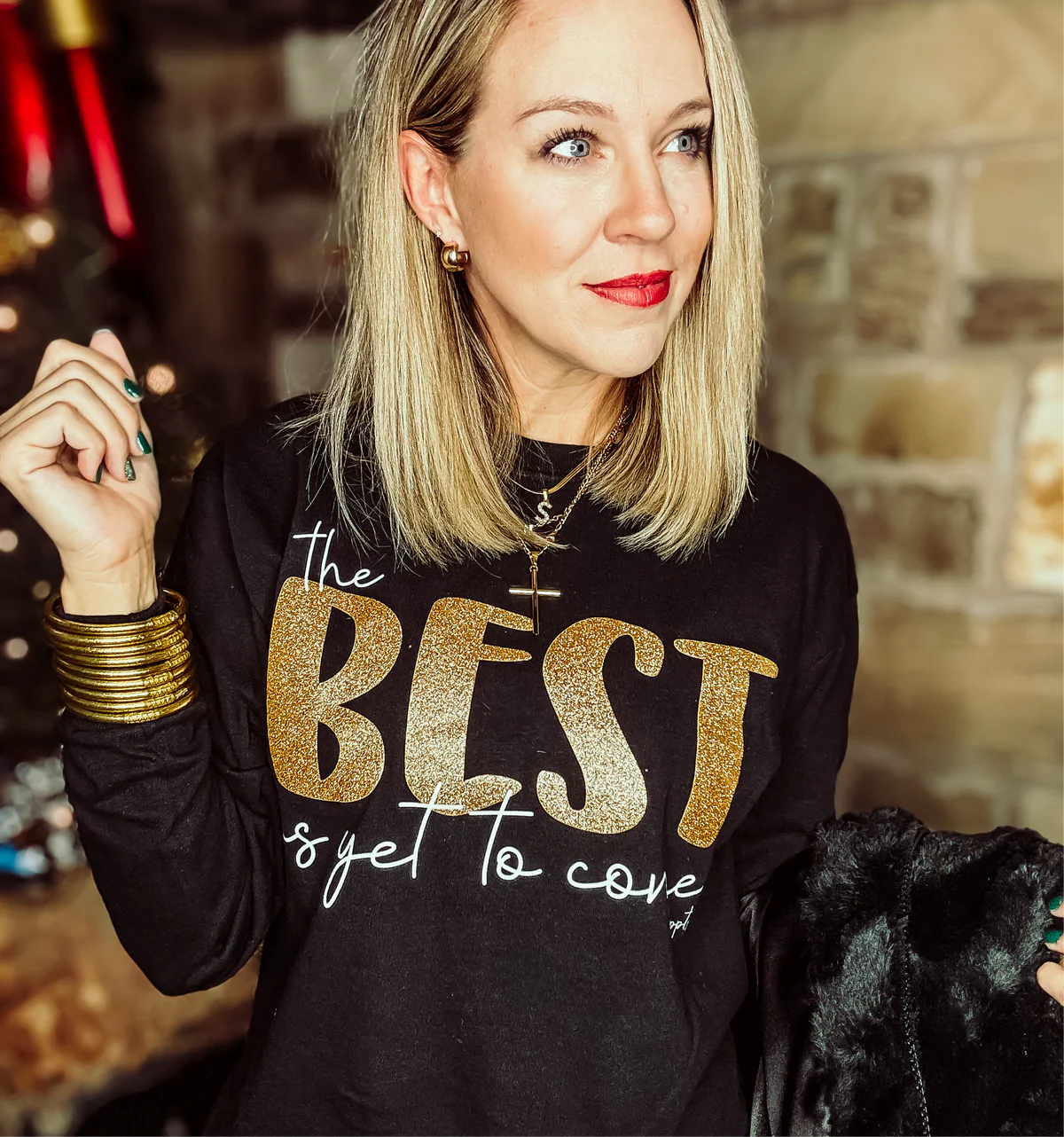 A woman wearing a black long sleeve graphic tee with the text 'THE BEST IS YET TO COME' in gold glitter.