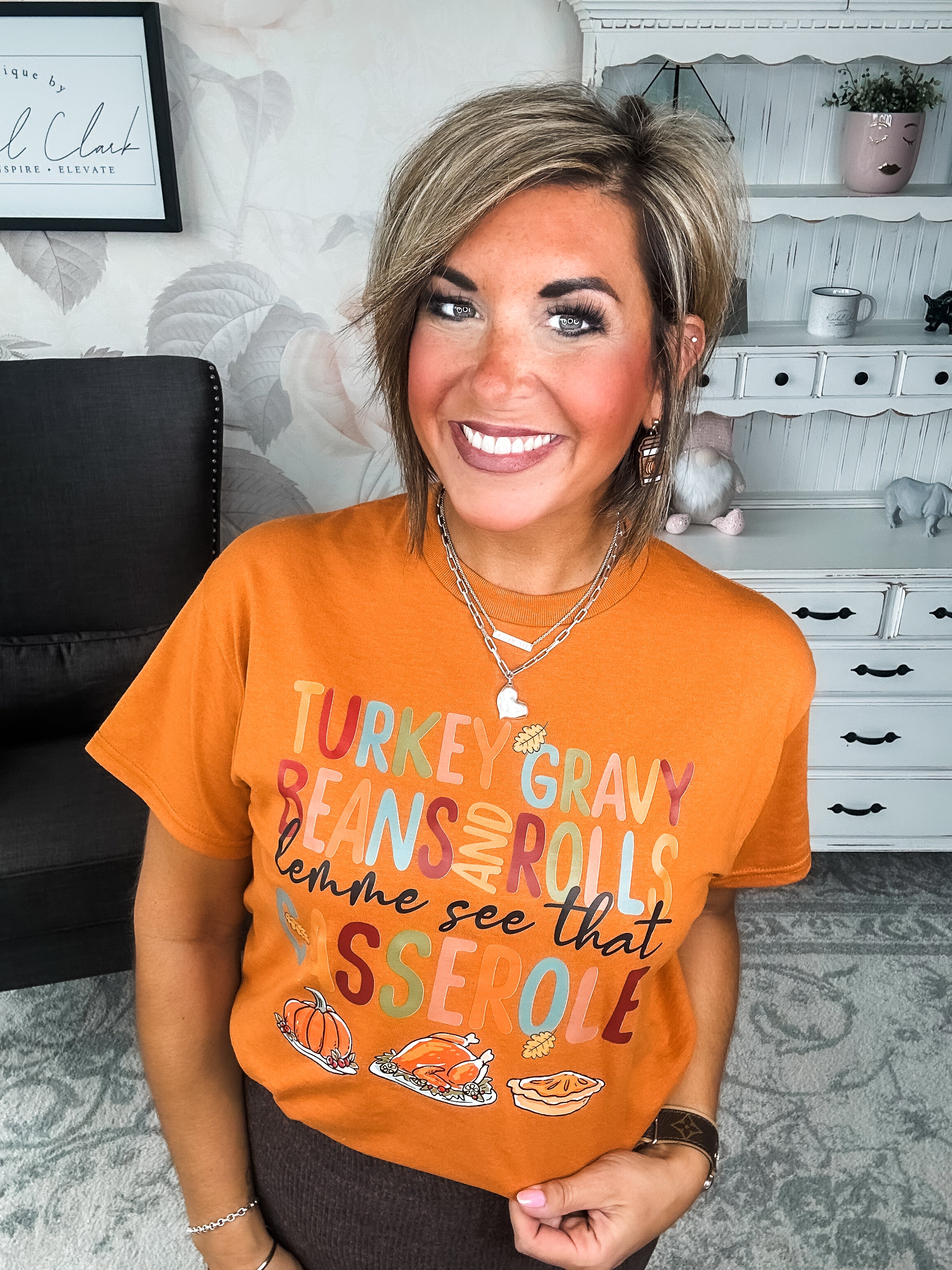 Woman wearing an orange t-shirt with Thanksgiving-themed text and graphics, that reads 'Turkey, gravy, beans and rolls, lemme see that casserole.'