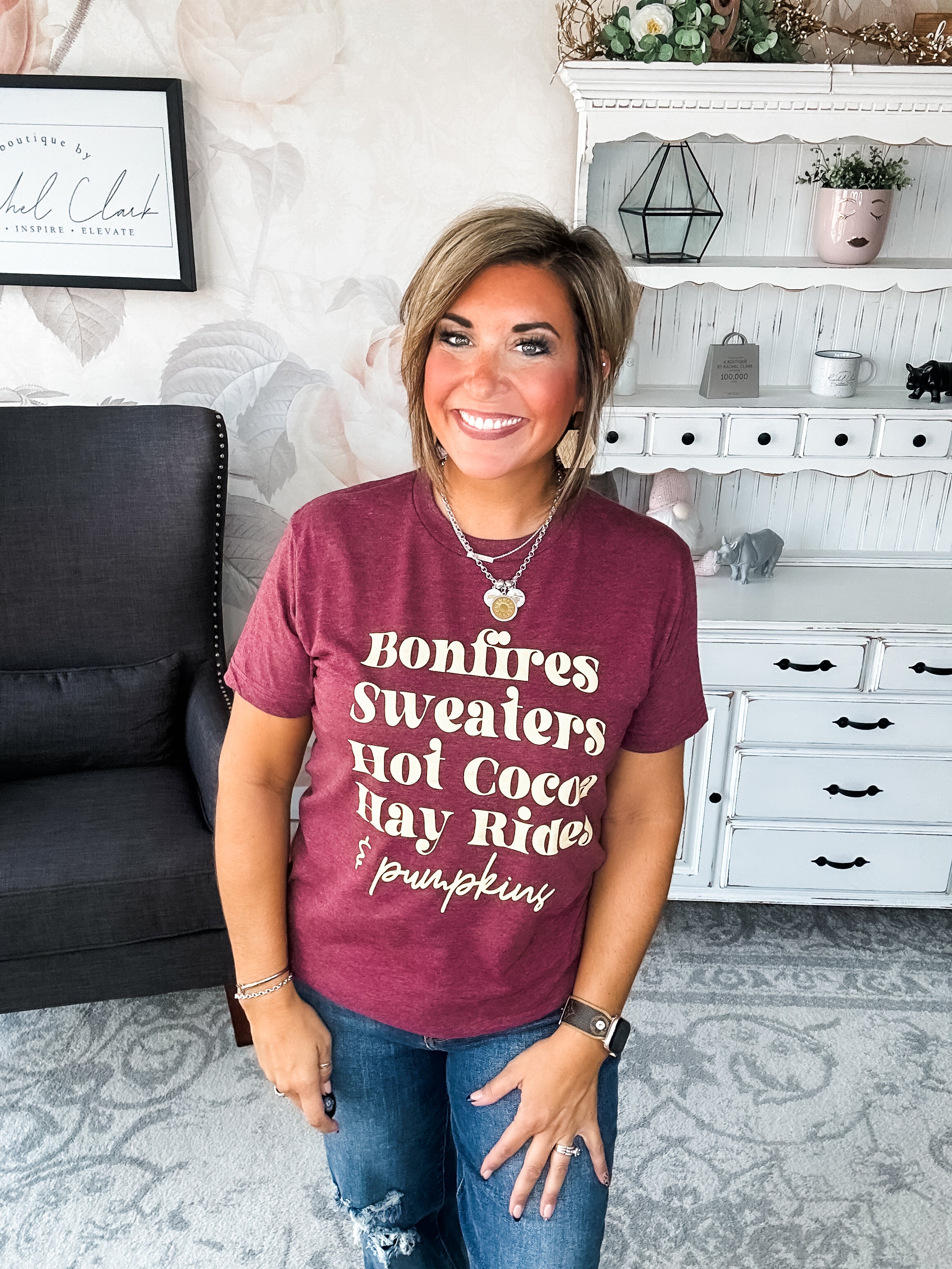 Woman wearing a maroon t-shirt that reads,  'Bonfires, Sweaters, Hot cocoa, Hay Rides and Pumpkins.' written in a cream text.