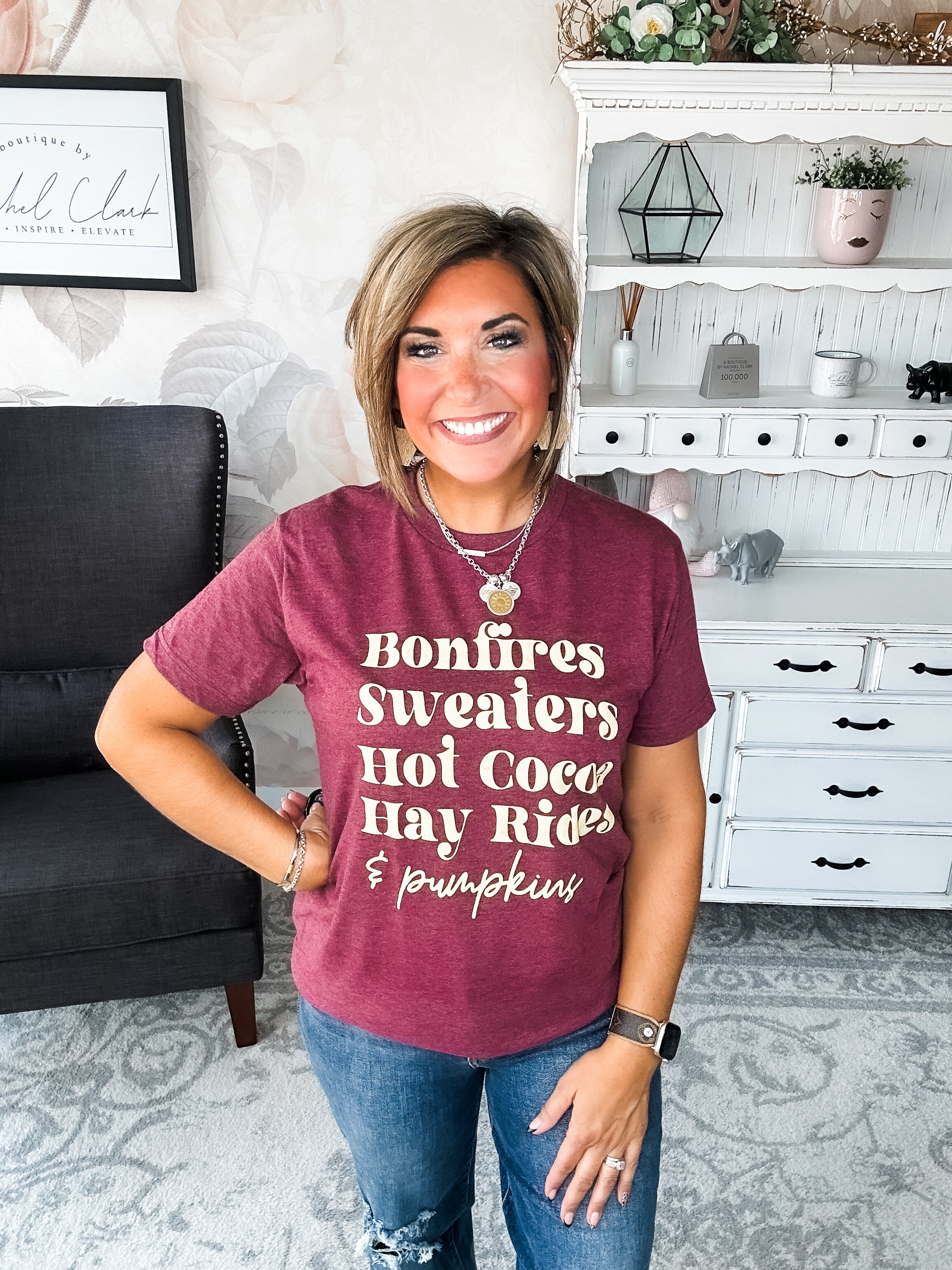 Woman wearing a maroon t-shirt that reads,  'Bonfires, Sweaters, Hot cocoa, Hay Rides and Pumpkins.' written in a cream text.