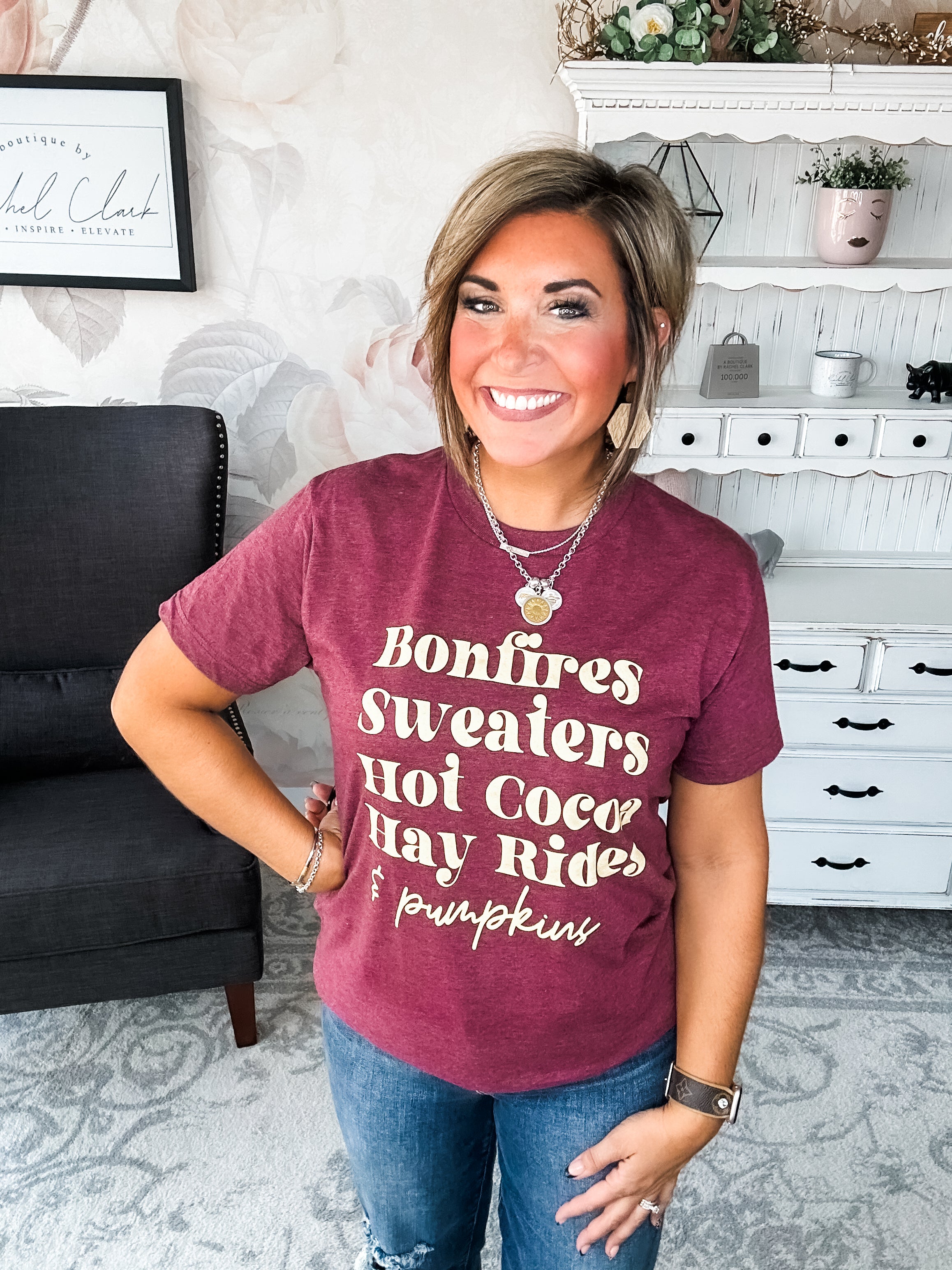 Woman wearing a maroon t-shirt that reads,  'Bonfires, Sweaters, Hot cocoa, Hay Rides and Pumpkins.' written in a cream text.