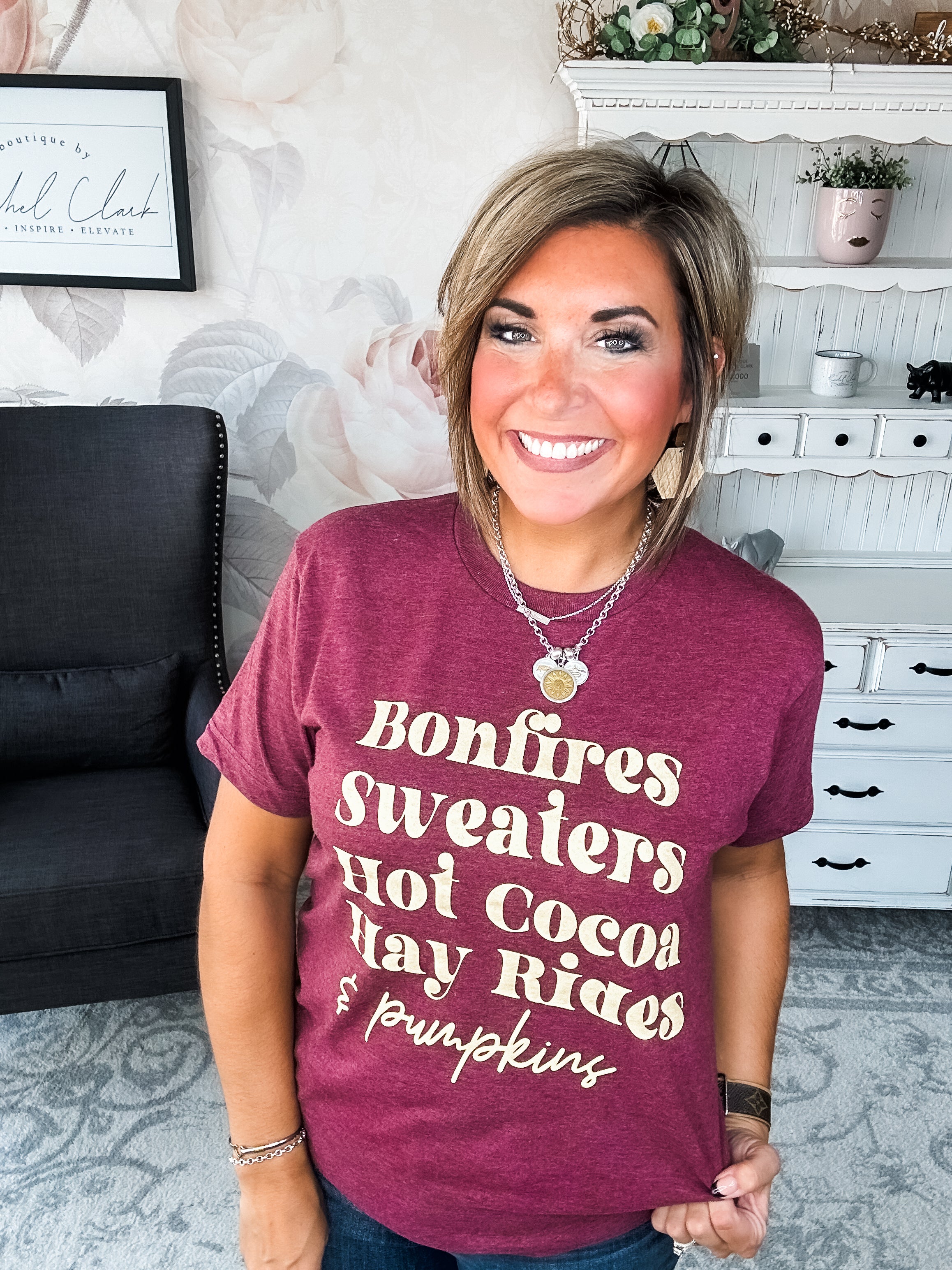 Woman wearing a maroon t-shirt that reads,  'Bonfires, Sweaters, Hot cocoa, Hay Rides and Pumpkins.' written in a cream text.