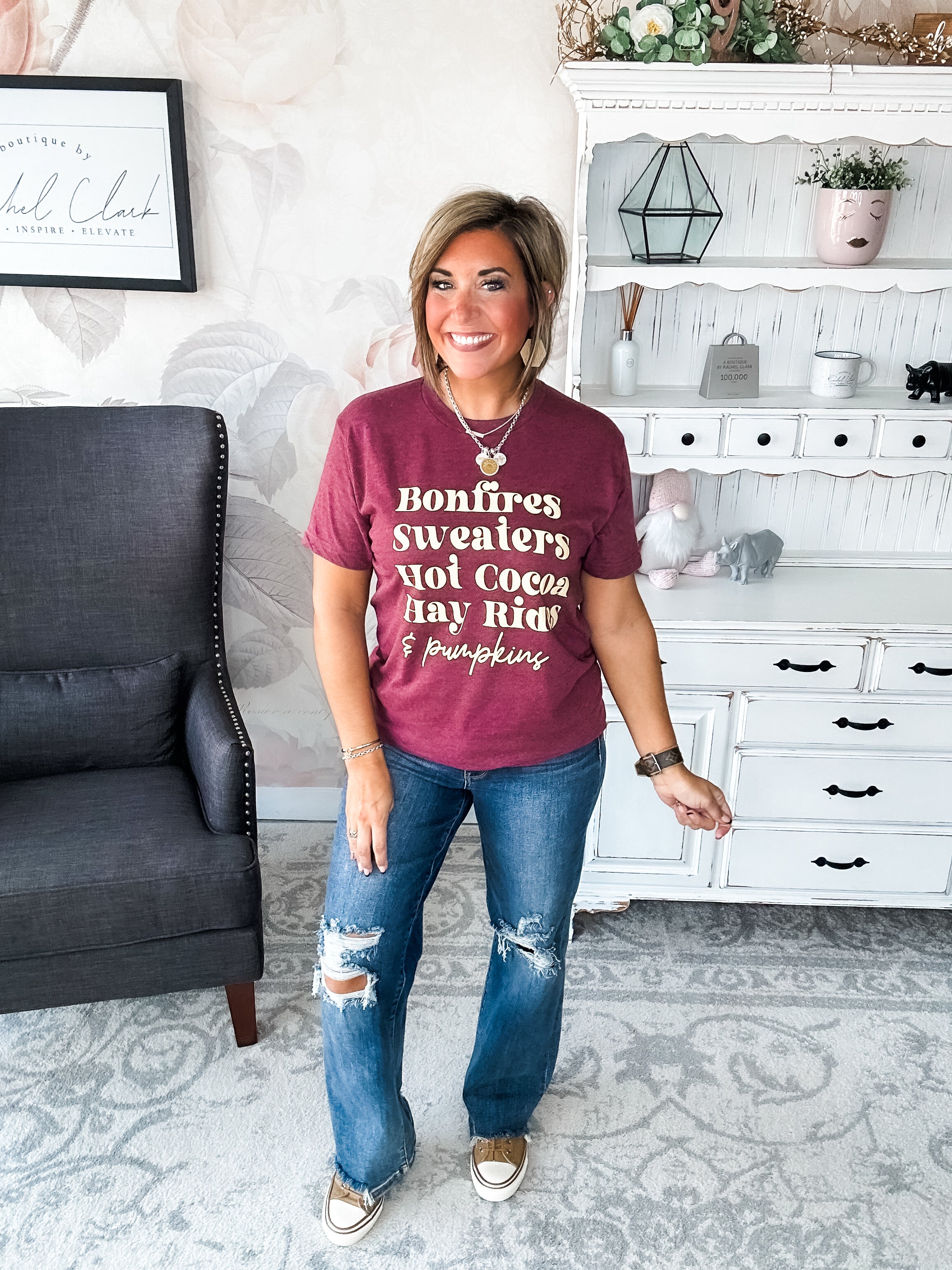 Woman wearing a maroon t-shirt that reads,  'Bonfires, Sweaters, Hot cocoa, Hay Rides and Pumpkins.' written in a cream text.