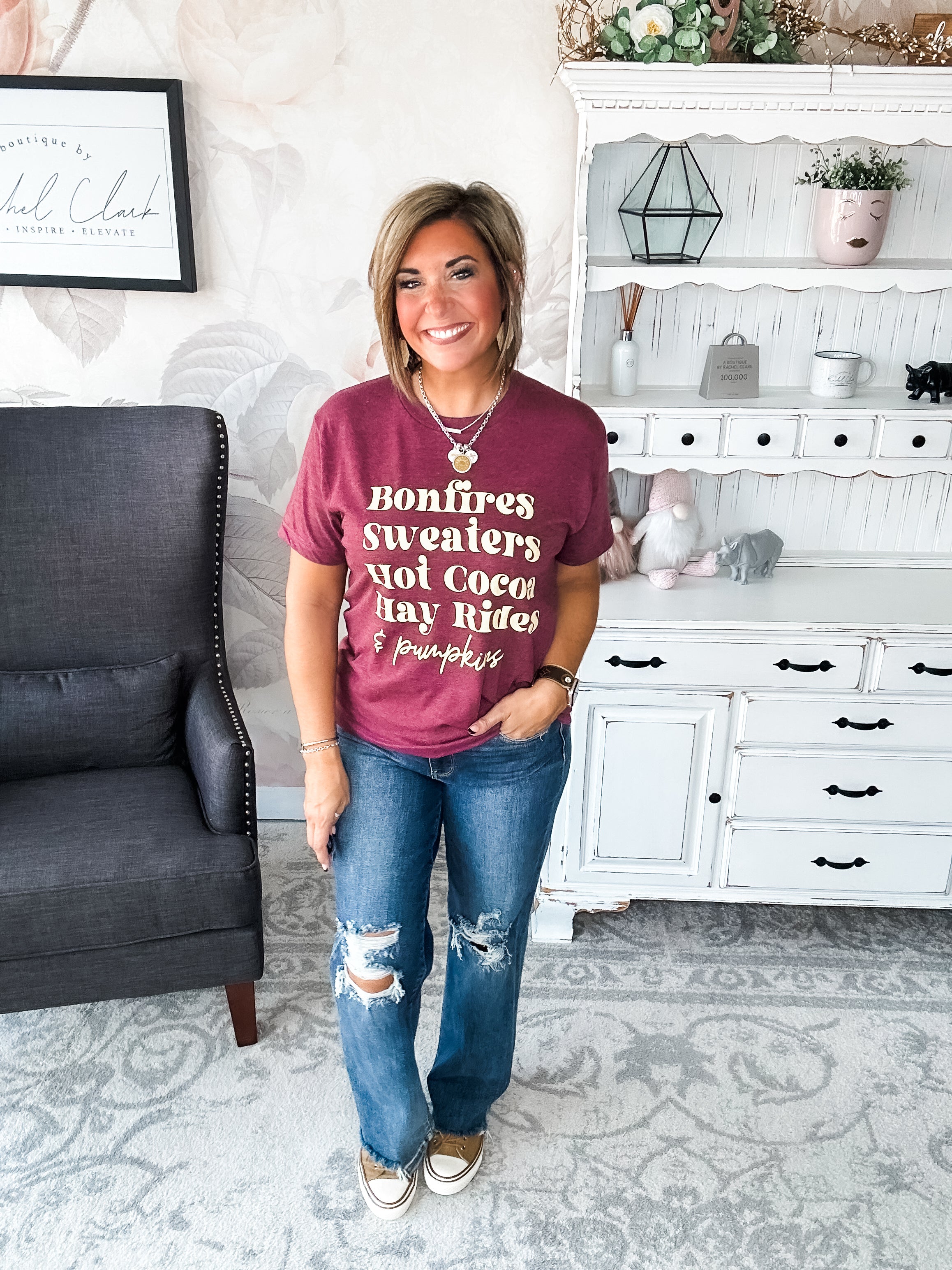 Woman wearing a maroon t-shirt that reads,  'Bonfires, Sweaters, Hot cocoa, Hay Rides and Pumpkins.' written in a cream text.