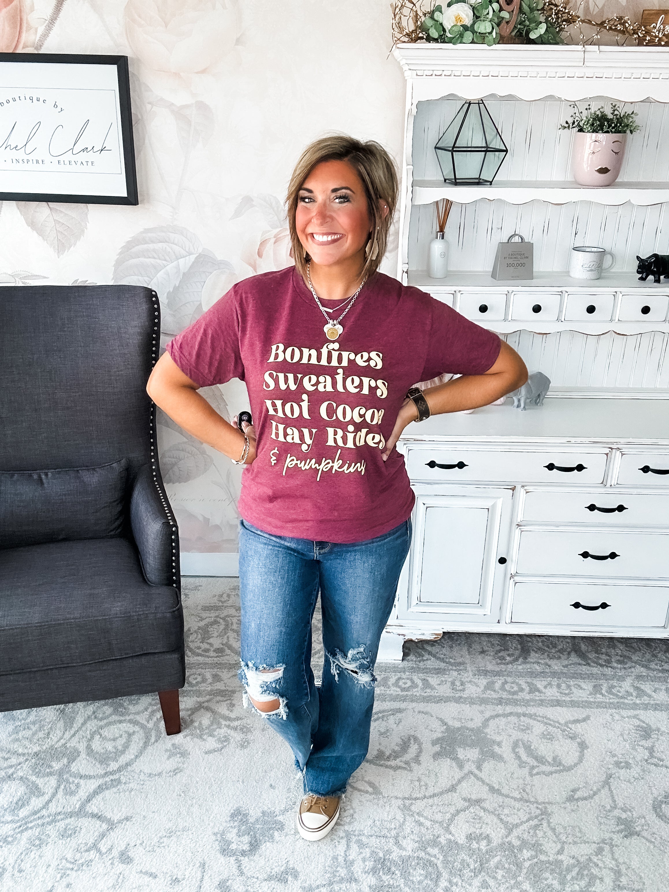 Woman wearing a maroon t-shirt that reads,  'Bonfires, Sweaters, Hot cocoa, Hay Rides and Pumpkins.' written in a cream text.