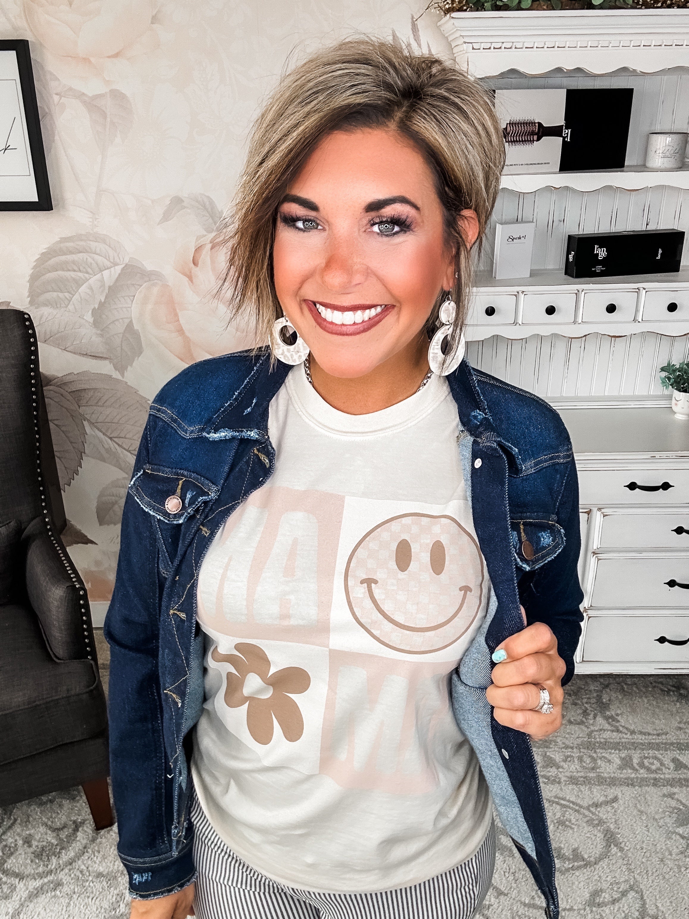 A woman wearing a cream crew-neck t-shirt with a smiley face graphic and text that reads 'MAMA'.
