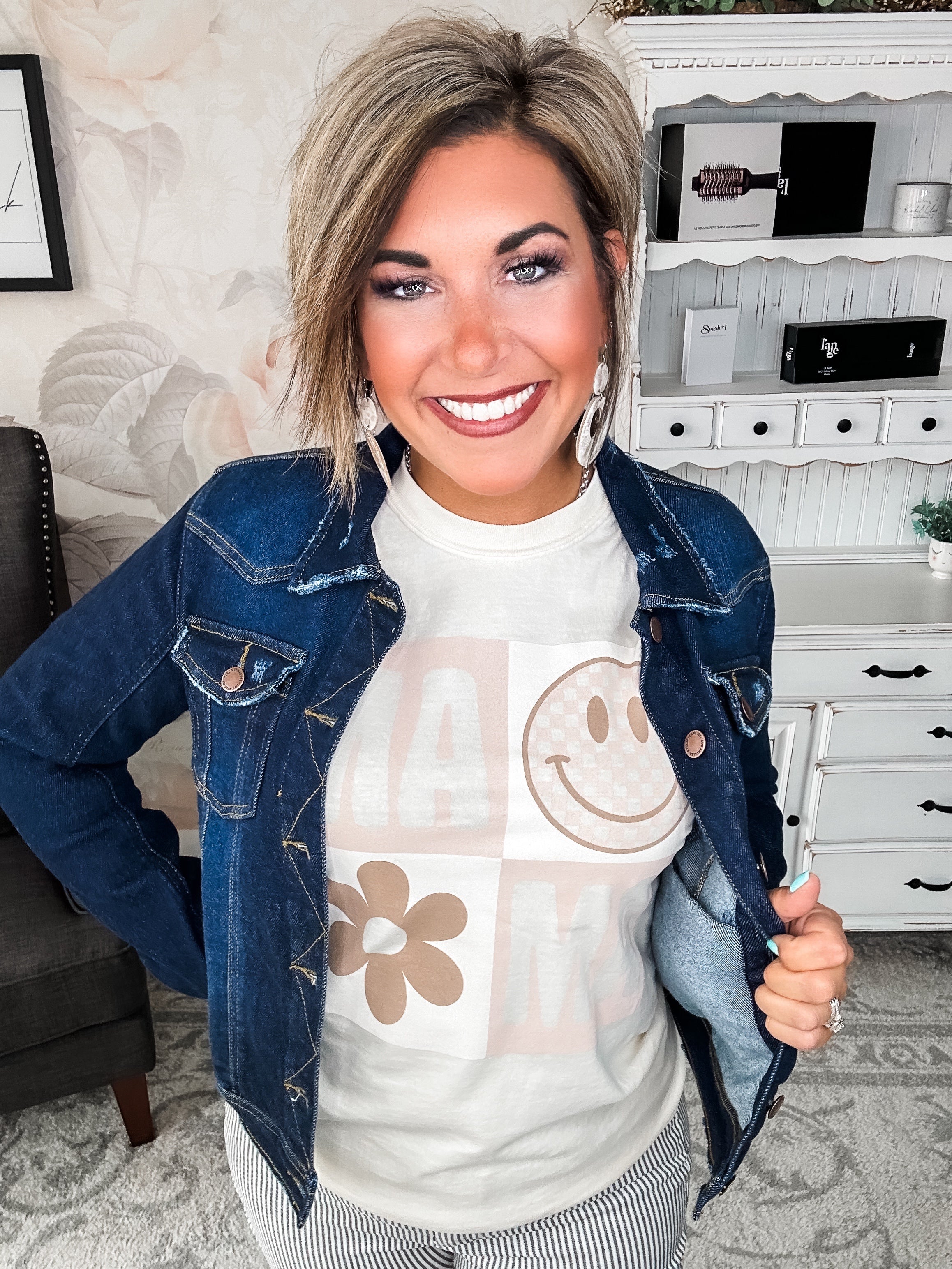 A woman wearing a cream crew-neck t-shirt with a smiley face graphic and text that reads 'MAMA'.