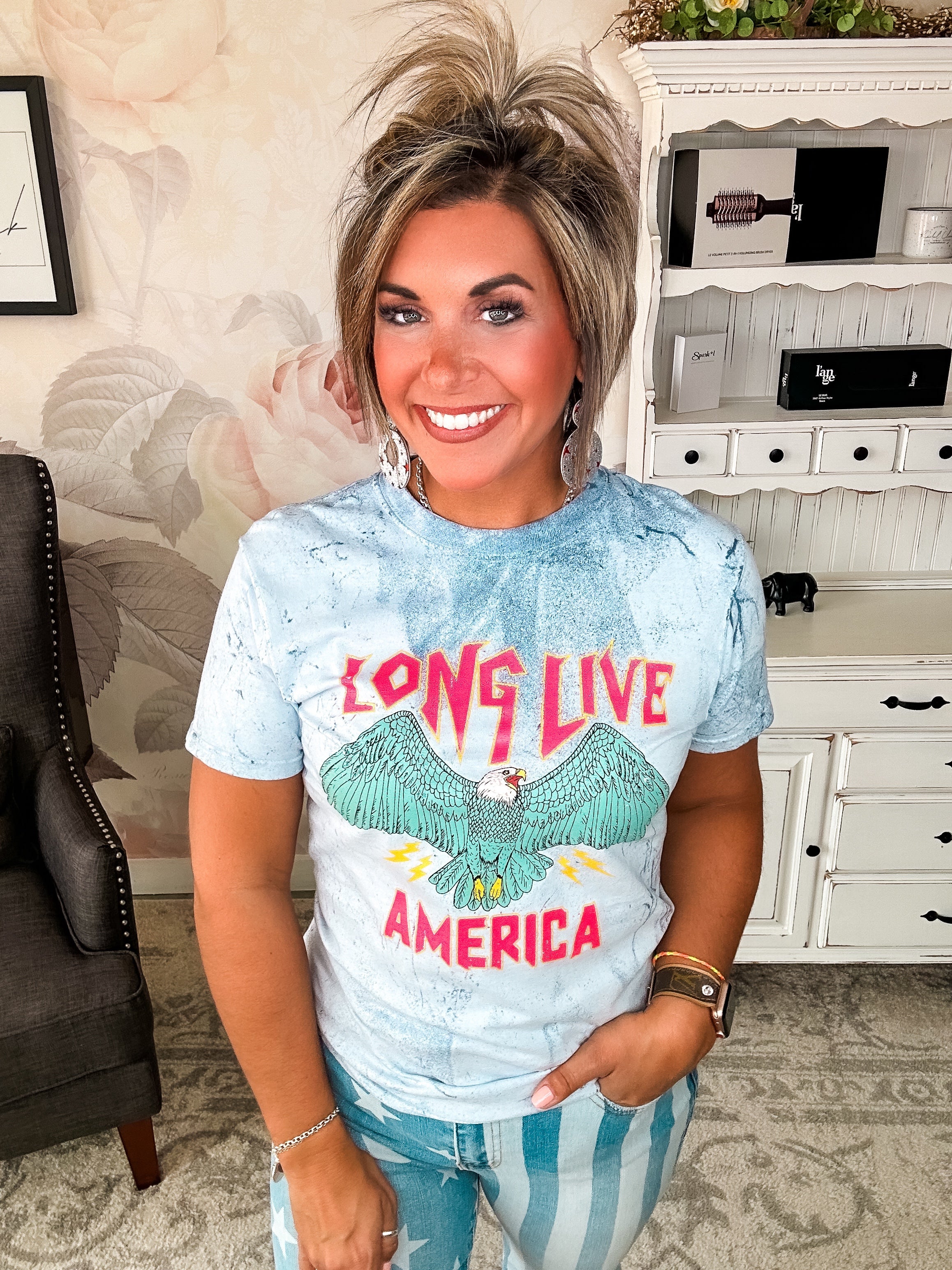 A person wearing a crew-neck, short-sleeve graphic tee with the phrase 'LONG LIVE AMERICA' printed on it, in a bleached blue color.