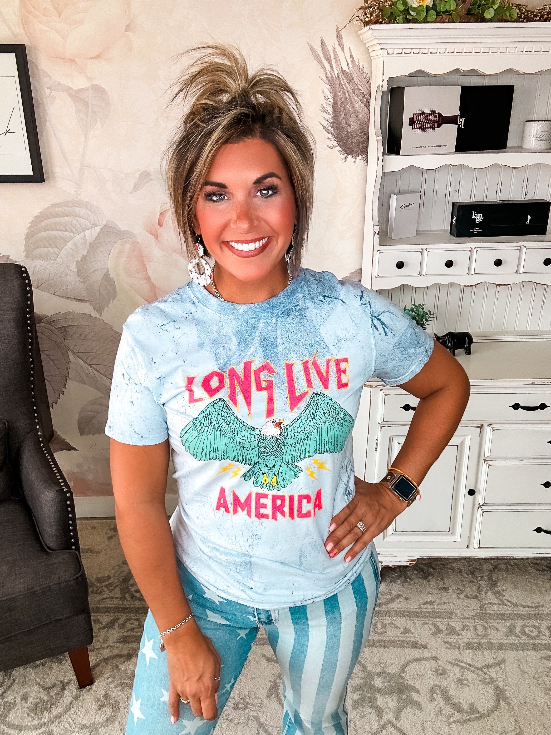 A person wearing a crew-neck, short-sleeve graphic tee with the phrase 'LONG LIVE AMERICA' printed on it, in a bleached blue color.