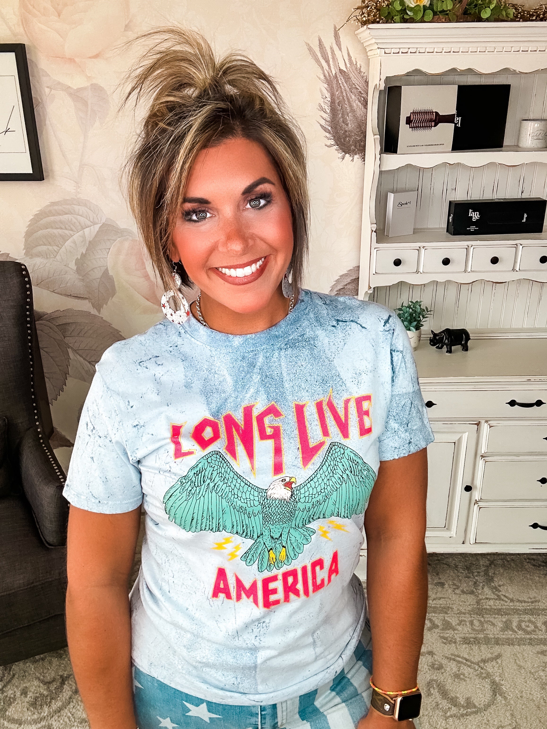 A person wearing a crew-neck, short-sleeve graphic tee with the phrase 'LONG LIVE AMERICA' printed on it, in a bleached blue color.