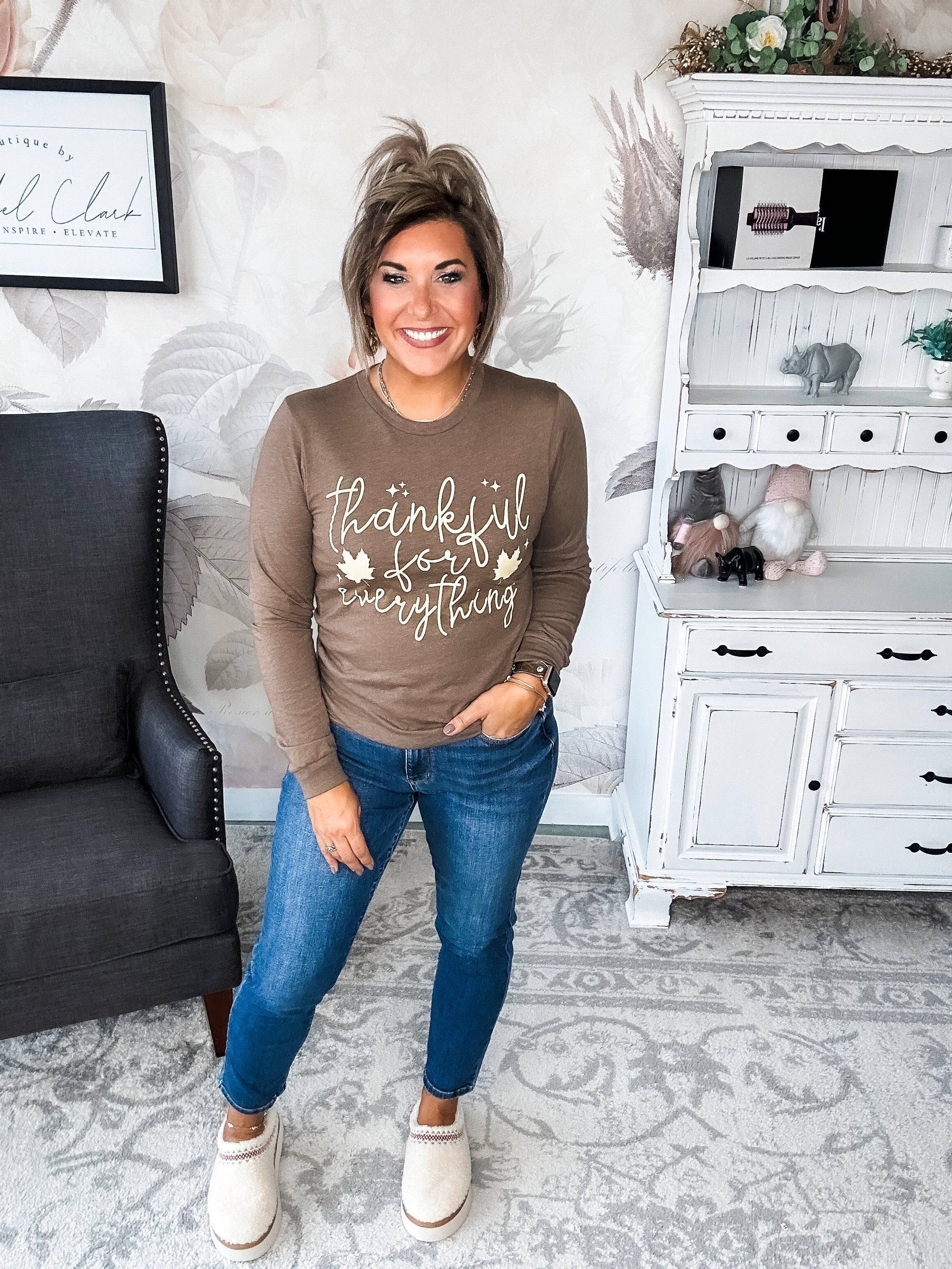 A woman wearing a brown long sleeve graphic tee that says 'thankful for everything' in pale yellow lettering.