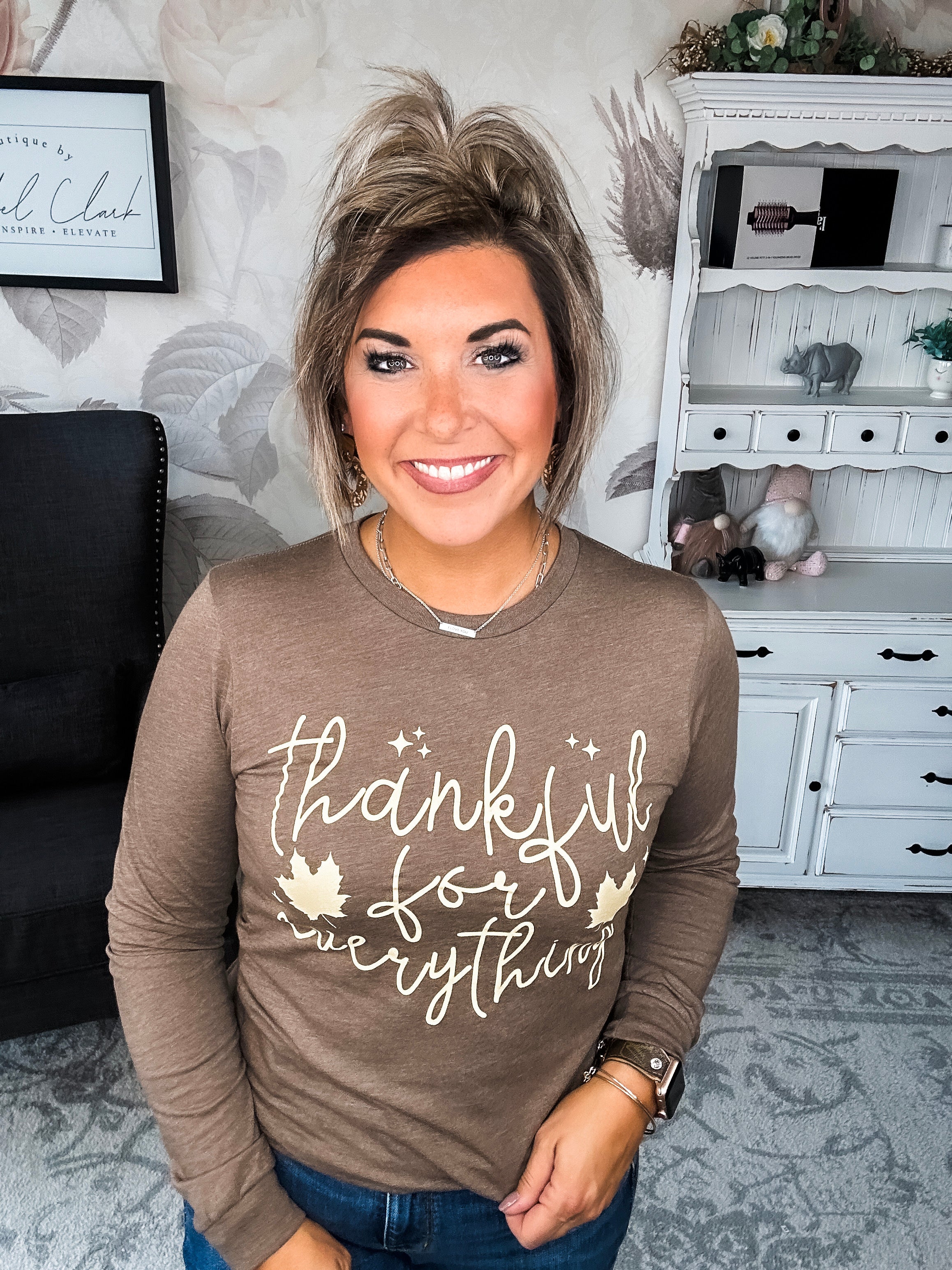 A woman wearing a brown long sleeve graphic tee that says 'thankful for everything' in pale yellow lettering.