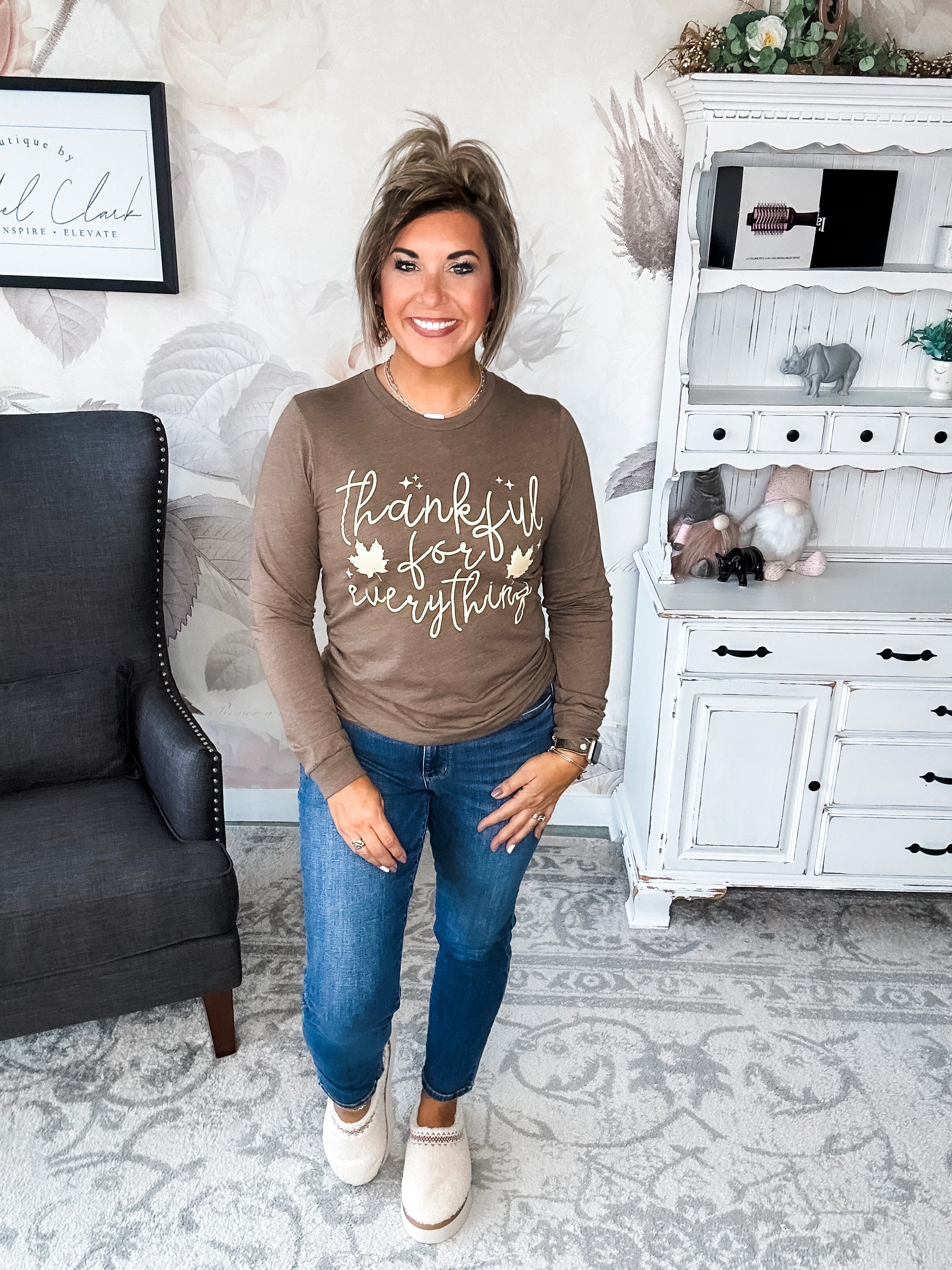A woman wearing a brown long sleeve graphic tee that says 'thankful for everything' in pale yellow lettering.