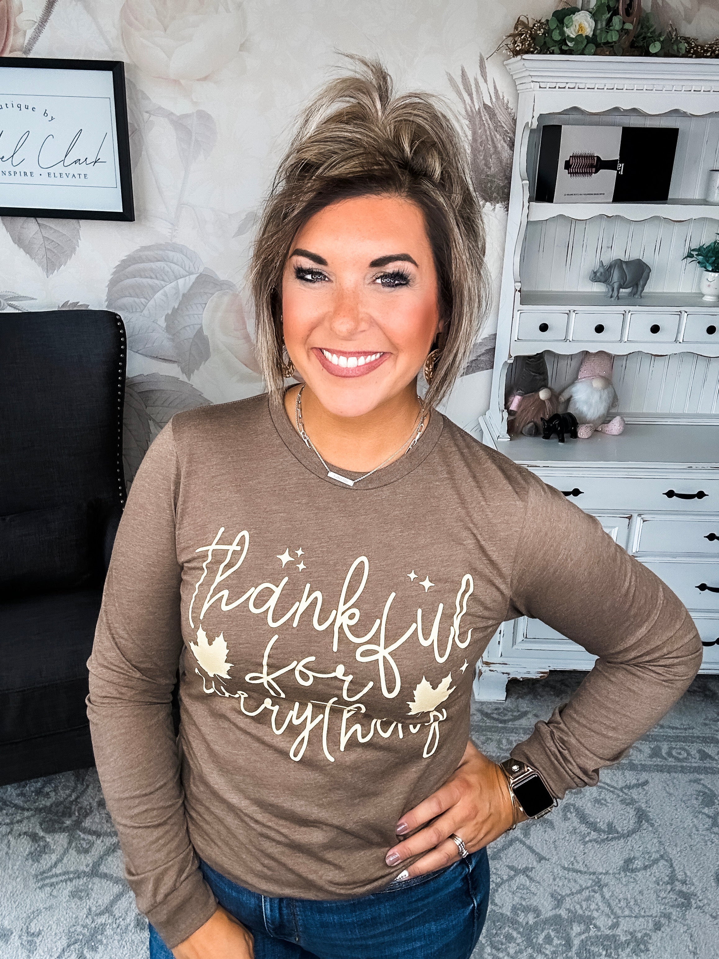 A woman wearing a brown long sleeve graphic tee that says 'thankful for everything' in pale yellow lettering.