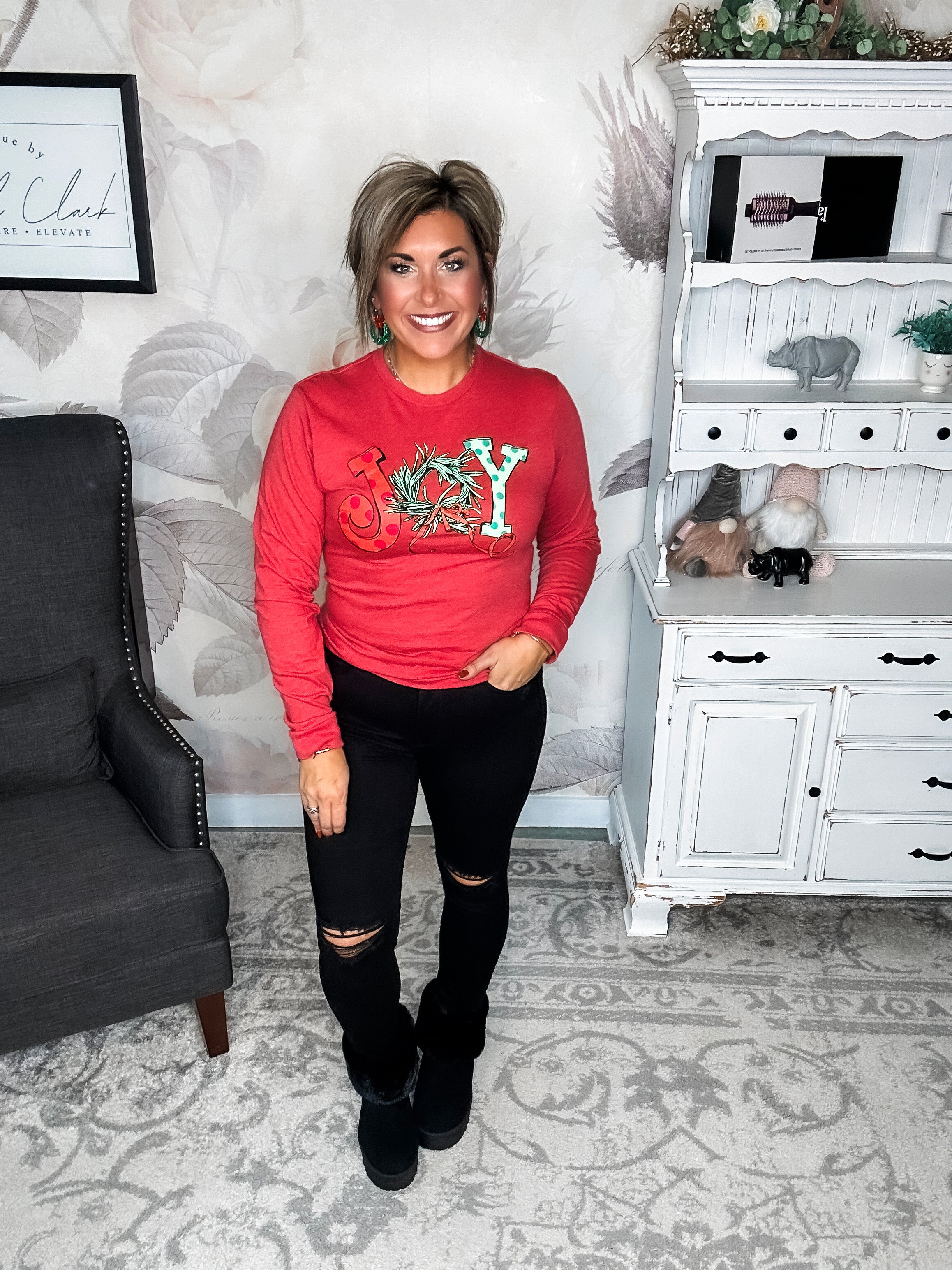 A woman wearing a red long sleeve graphic tee with the word 'JOY' printed on the front with Christmas graphics.