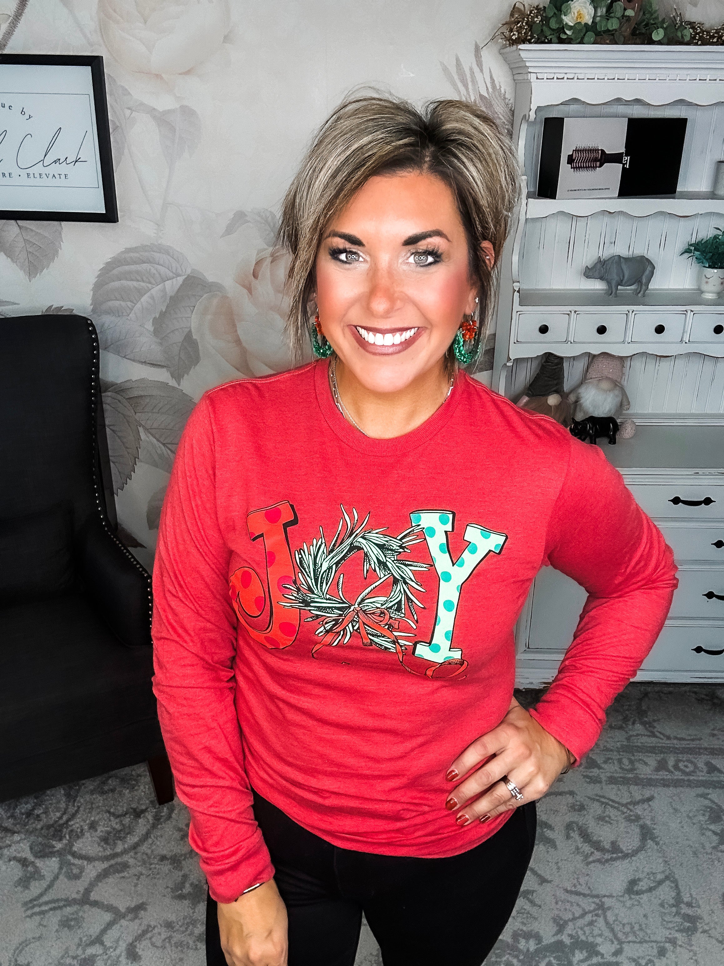 A woman wearing a red long sleeve graphic tee with the word 'JOY' printed on the front with Christmas graphics.