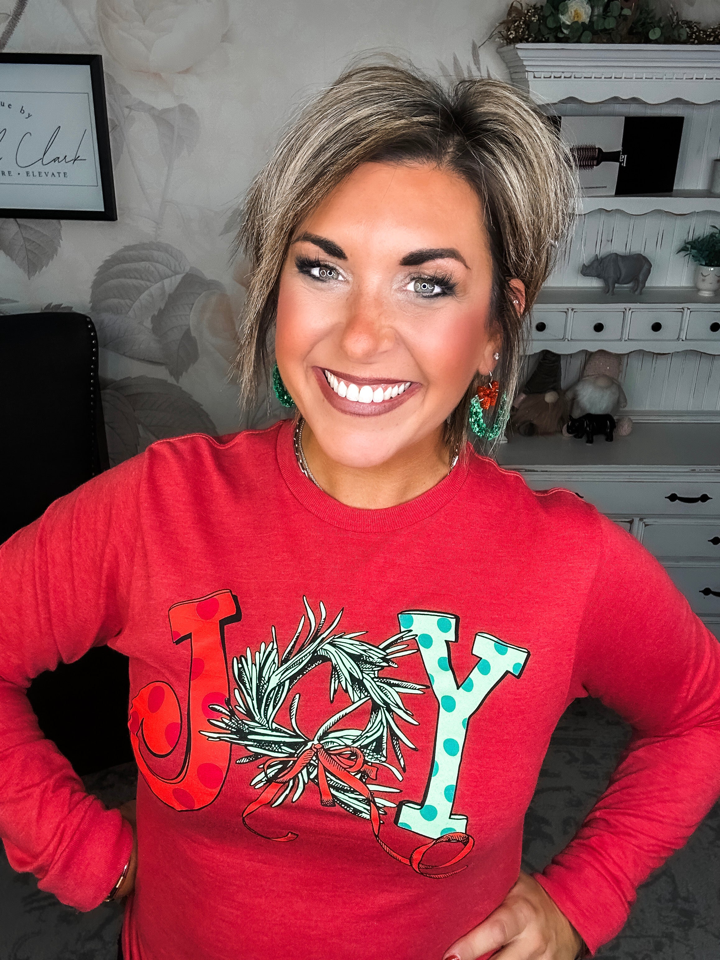 A woman wearing a red long sleeve graphic tee with the word 'JOY' printed on the front with Christmas graphics.