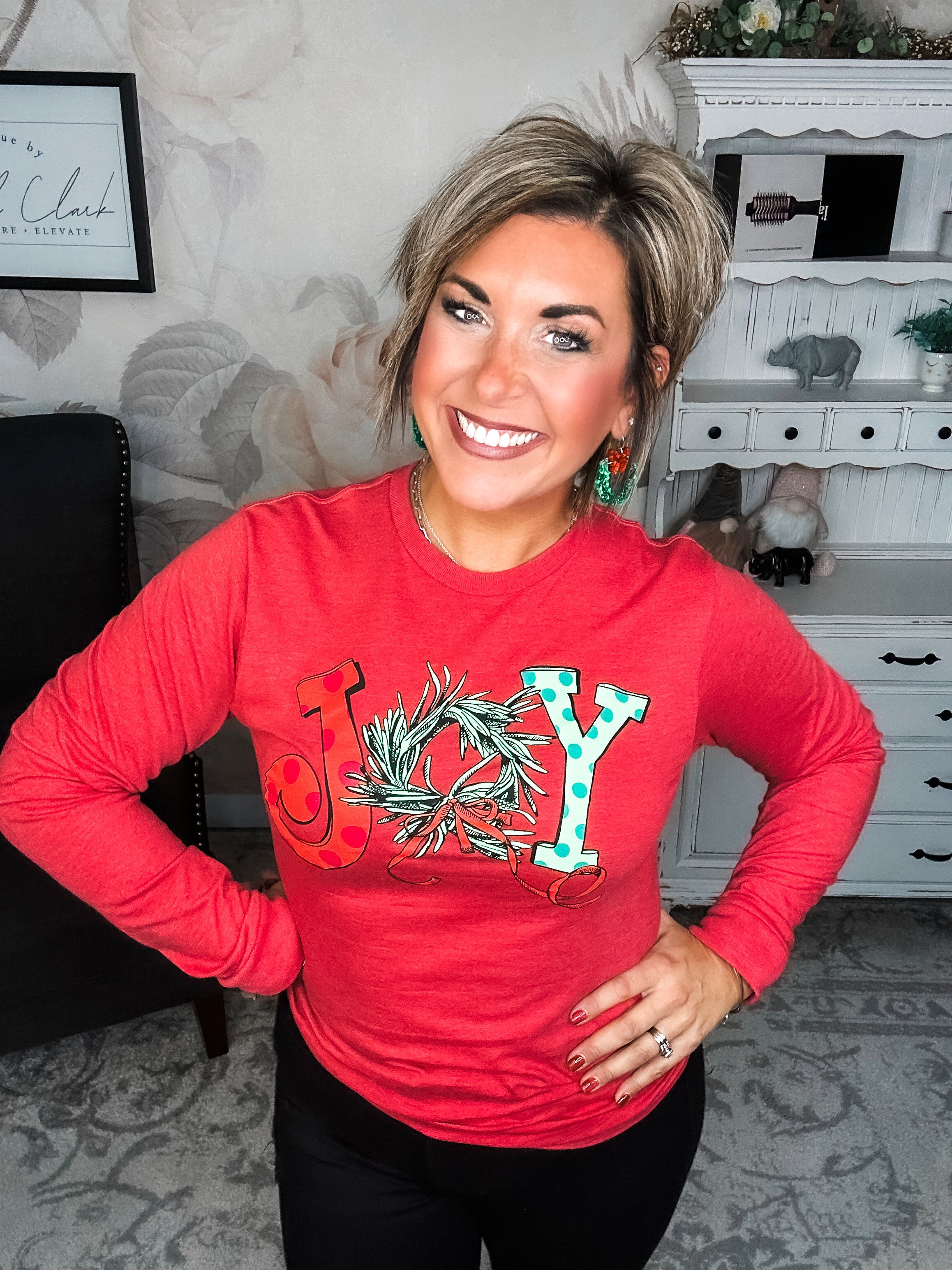 A woman wearing a red long sleeve graphic tee with the word 'JOY' printed on the front with Christmas graphics.