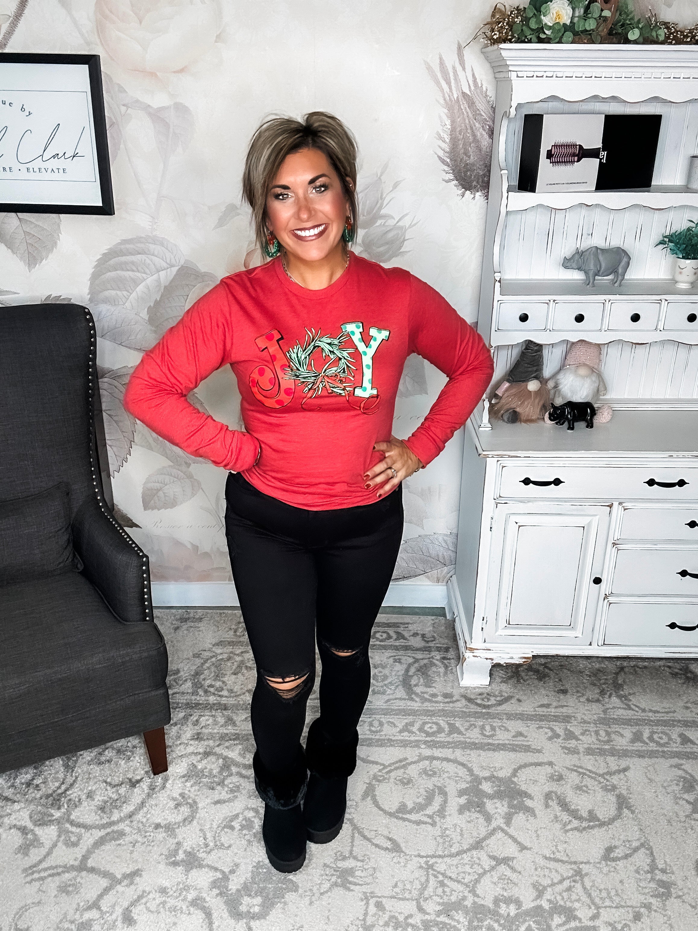 A woman wearing a red long sleeve graphic tee with the word 'JOY' printed on the front with Christmas graphics.