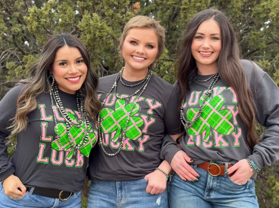 three people wearing a dark grey graphic tee with a color combination of pink, green, and white ink plaid, that reads 'Lucky Lucky Lucky'
