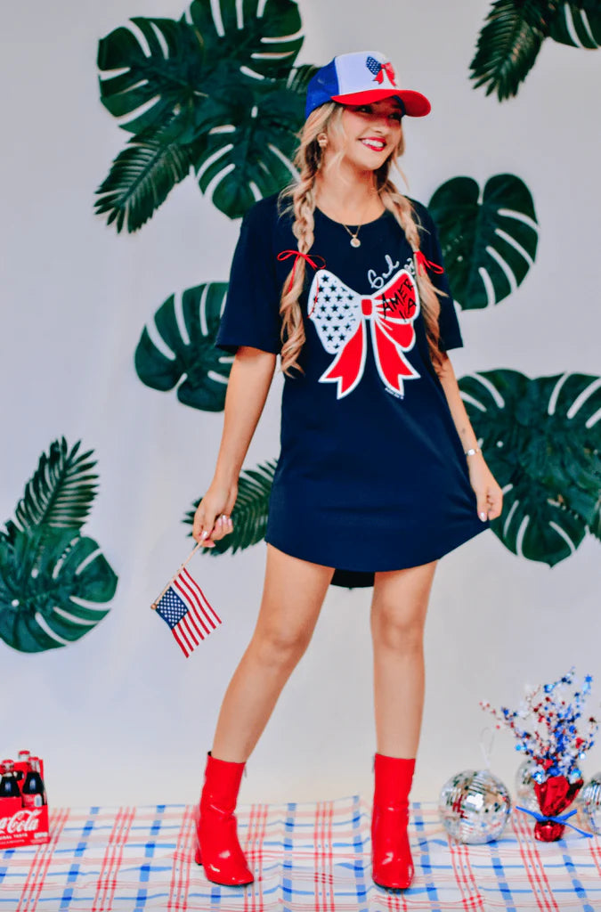 A woman wearing a navy t-shirt dress with a large bow graphic on the front and small text that reads 'God Bless America'.