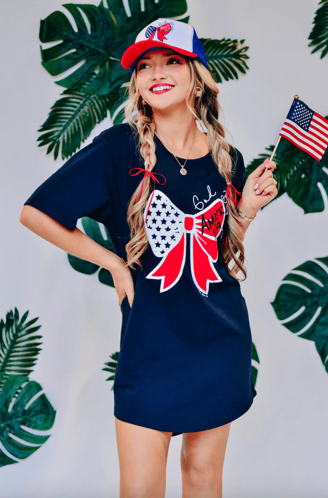 A woman wearing a navy t-shirt dress with a large bow graphic on the front and small text that reads 'God Bless America'.