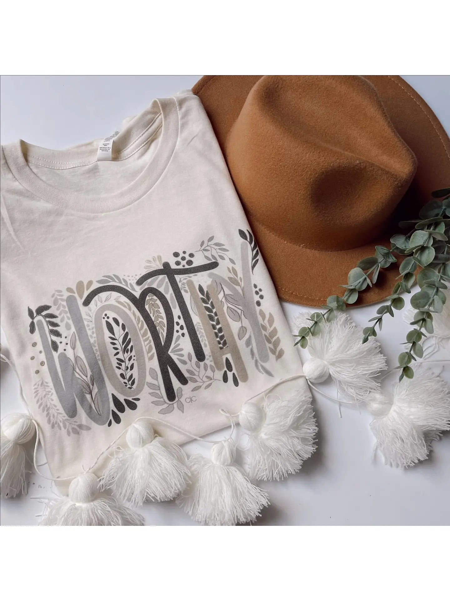A cream graphic tee with the word 'worthy' displayed in a floral style font.