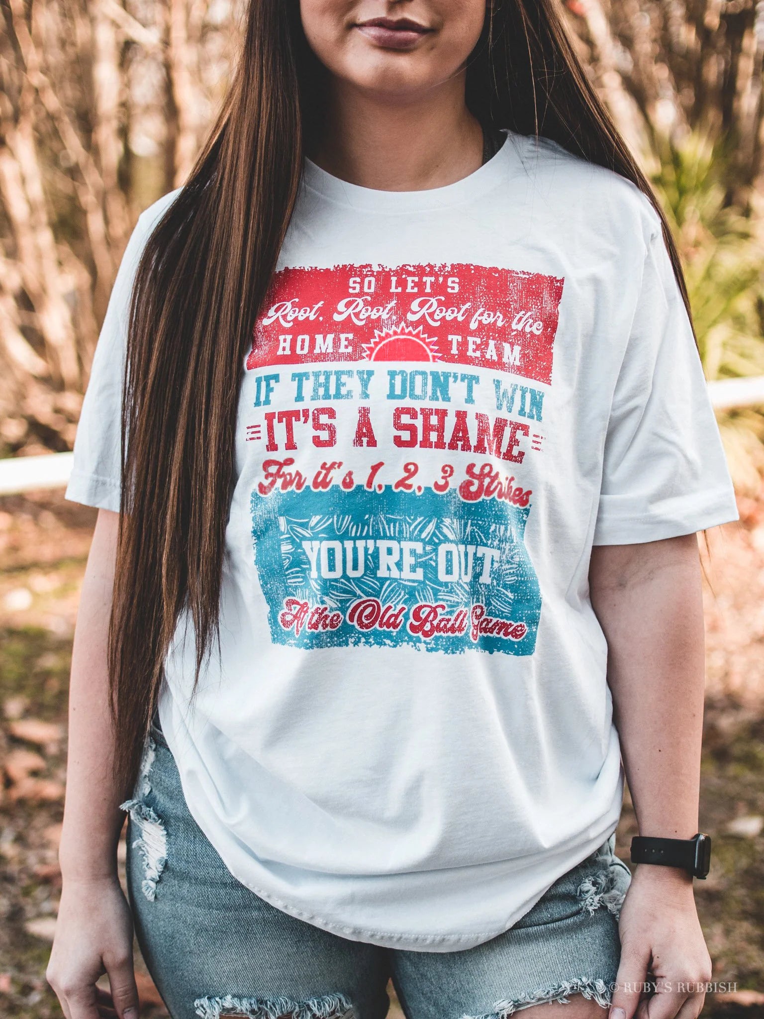 A woman wearing a white graphic tee with red and blue text and graphics, that says 'So let's root, root, root for the home team. If they don't win it's a shame. For it's 1,2,3 strikes you're out at the old ball game'