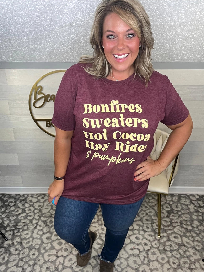 Woman wearing a maroon t-shirt that reads,  'Bonfires, Sweaters, Hot cocoa, Hay Rides and Pumpkins.' written in a cream text. 