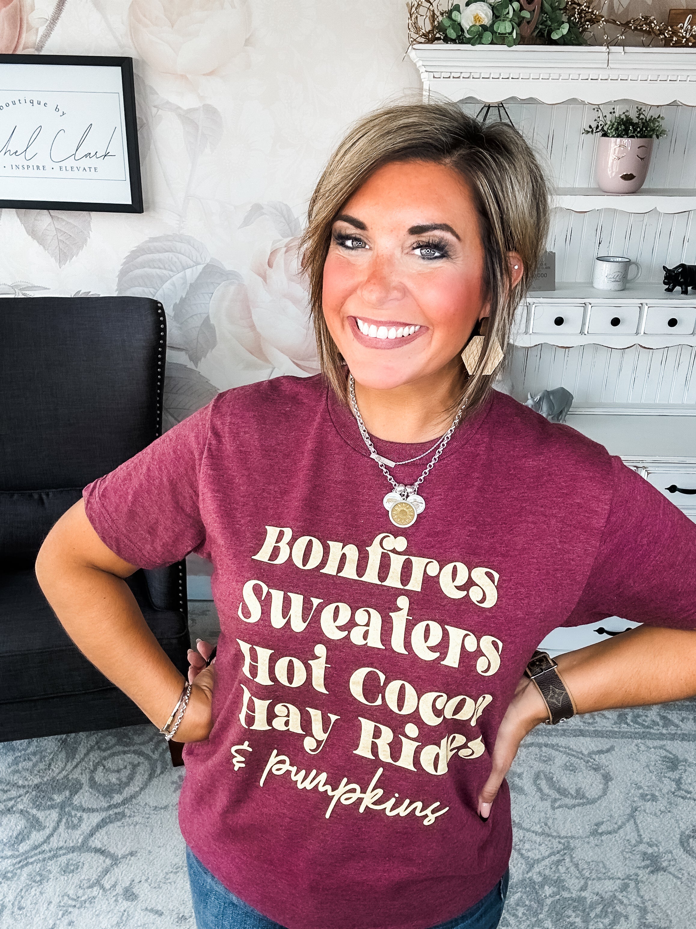 Woman wearing a maroon t-shirt that reads,  'Bonfires, Sweaters, Hot cocoa, Hay Rides and Pumpkins.' written in a cream text. 