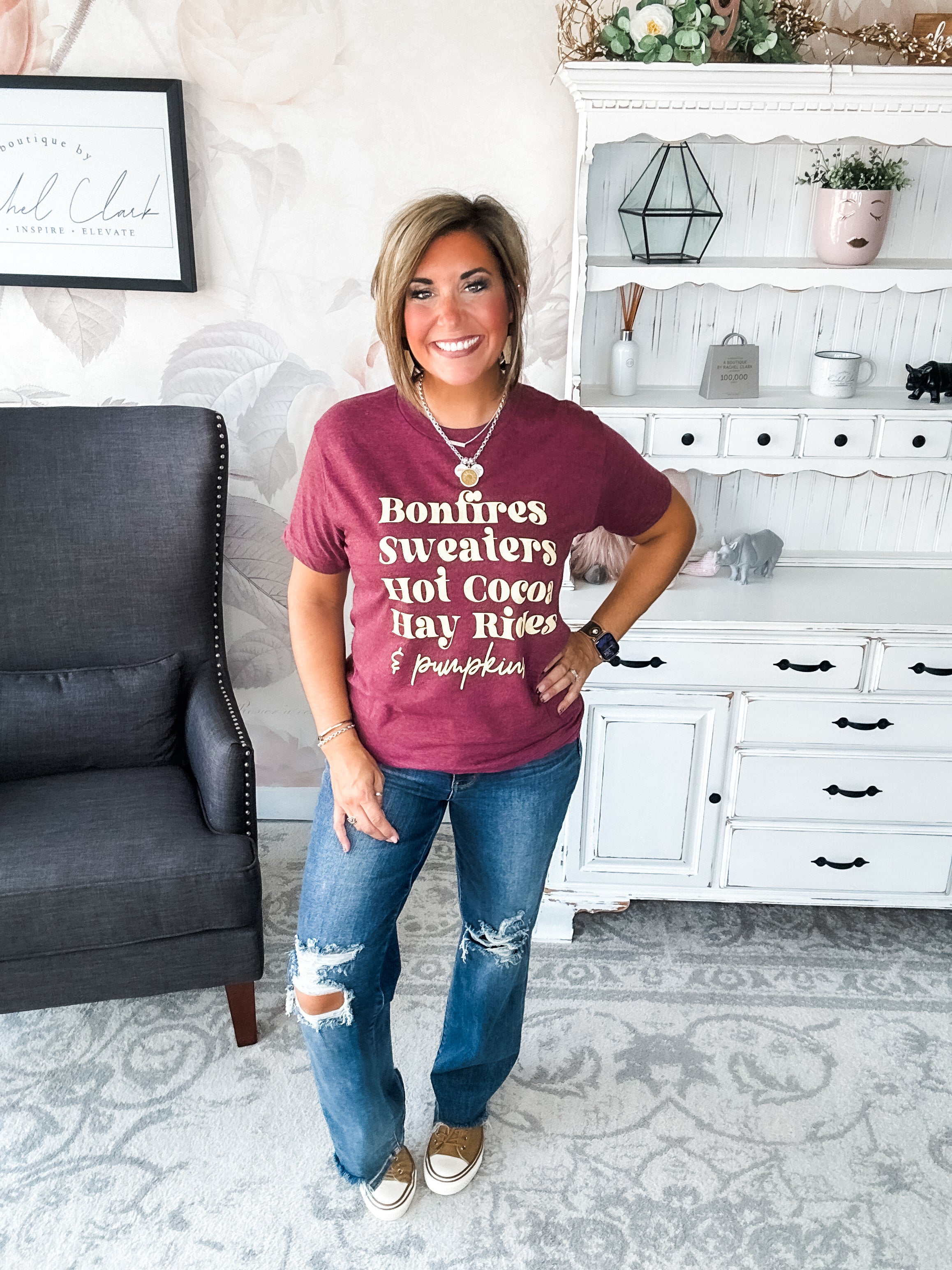 Woman wearing a maroon t-shirt that reads,  'Bonfires, Sweaters, Hot cocoa, Hay Rides and Pumpkins.' written in a cream text. 